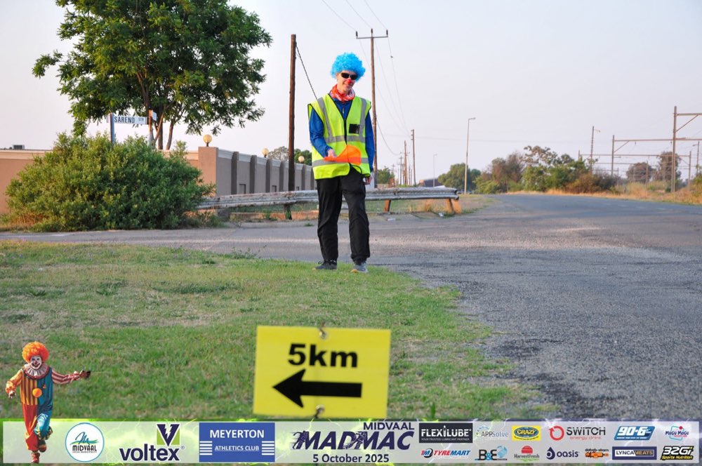 A course marshal in a blue wig and hi-vis vest at the 5km marker on a Randvaal road