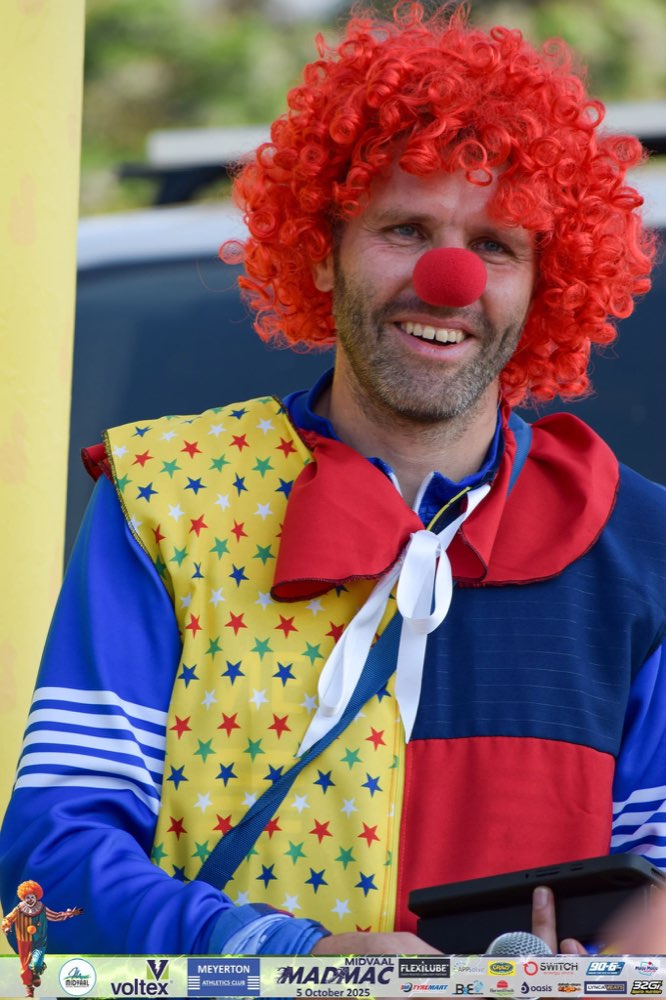 The race MC in full clown costume with a red wig, holding a tablet and microphone